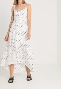 Bílé maxi šaty s puntíky, tenkými ramínky, zapínáním na knoflíky vpředu a asymetrickým lemem. Nosí se s černými skluzovými sandály.