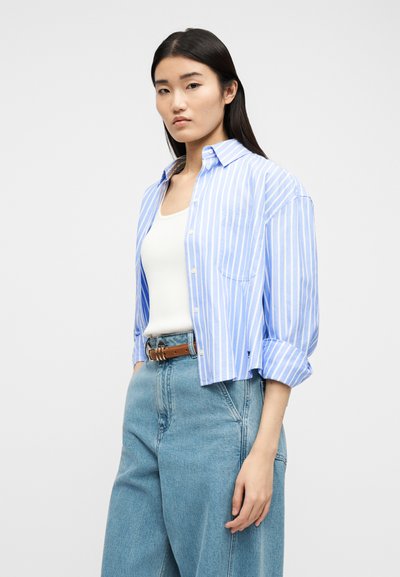 Νέα γυναίκα με μακριά σκούρα μαλλιά που φορούν ένα ανοιχτό μπλε και άσπρο ριγέ πουκάμισο πάνω από ένα λευκό αμάνικο τοπ, τζιν ψηλόμεσο και καφέ ζώνη.