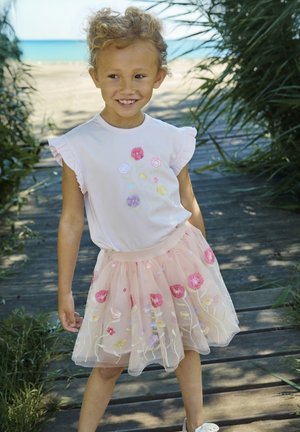 Jong meisje dat glimlacht, draagt een lichtroze jurk met bloemenborduursels, staand op een houten pad nabij een strand omgeven door groen.
