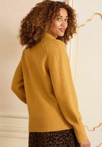 Mustár sárga kötött pulóver bordás szegéllyel és finom levélmintával a vállakon, hosszú ujjakkal és laza szabással.