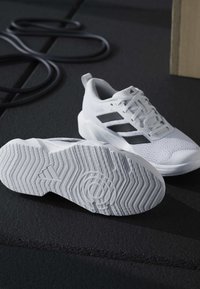 Λευκά αθλητικά παπούτσια Adidas με μαύρες ρίγες, το ένα δείχνει την υφή της σόλας, τοποθετημένα πάνω σε σκοτεινό στρώμα άσκησης κοντά σε λαστιχένια ταινία αντίστασης.