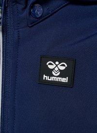 En marinblå jacka i tyg har en svart rektangulär logotypatch med ett vitt emblem och texten "hummel", accentuerad av en blå knapp.