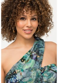 Robe asymétrique à une épaule en tissu léger ornée d'un imprimé coloré de papillons dans des tons de vert, bleu et violet. Grandes boucles d'oreilles en or.