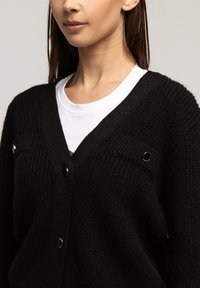 Czarny kardigan z teksturowym splotem, z dekoltem w serek, zapięciem na napy i dwoma przednimi kieszeniami. Udekorowany czarnymi detalami wykończeniowymi.