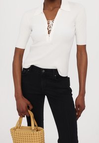 Vit ribbad topp med V-ringning, korta ärmar och snörningsdetalj fram, matchad med svarta jeans och en liten vävd gul väska.