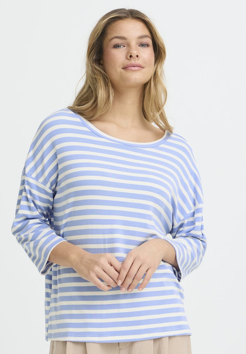 Jeune femme aux cheveux blonds portant un haut à manches longues rayé bleu et blanc, les mains jointes devant elle sur un fond blanc.