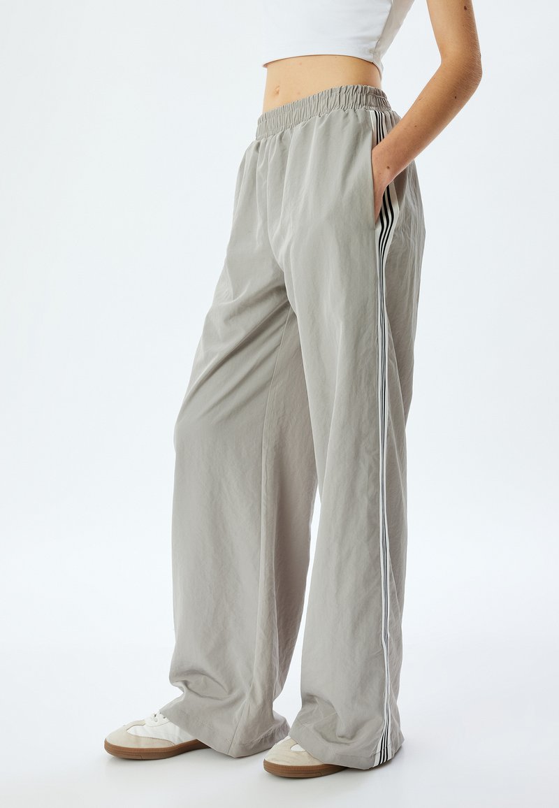 Pantalons larges gris clair avec une taille élastique, des poches latérales et des rayures verticales noires le long des coutures extérieures. Design texturé et décontracté.