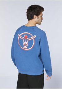 Blauer Sweatshirt mit einem großen rot-weißen aufgestickten Motiv auf der Rückseite, das eine Figur mit ausgebreiteten Armen in einem Kreis darstellt.