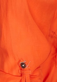 Tissu orange avec une texture lisse, présente un bouton marron et des détails de couture. Le design inclut une section froncée avec un cordon de serrage.