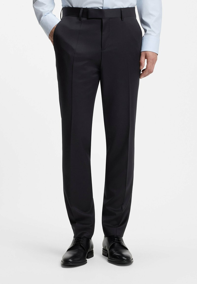 Homme portant un pantalon habillé noir avec une ceinture, une chemise bleu clair et des chaussures en cuir noires, debout, les mains partiellement dans les poches.