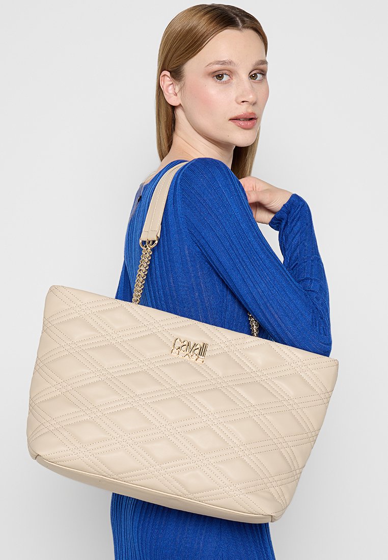 Bolso tote acolchado en color beige con correa de cadena y un acento de logo dorado. Textura de cuero suave, forma rectangular y diseño espacioso.
