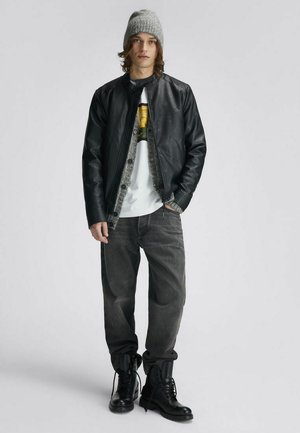 Giovane che indossa un berretto di lana grigio, giacca di pelle nera, cardigan grigio, t-shirt bianca con stampa, jeans neri sbiaditi e stivali neri da combattimento.