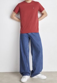 Rød bomulds t-shirt med korte ærmer, lille logo på brystet; parret med løstsiddende blå denim jeans og hvide sneakers.