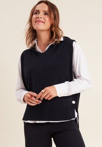 Pull sans manches en tricot marine avec un col en V, tissu texturé, fentes latérales et accents de boutons. Superposé sur une chemise blanche à col.