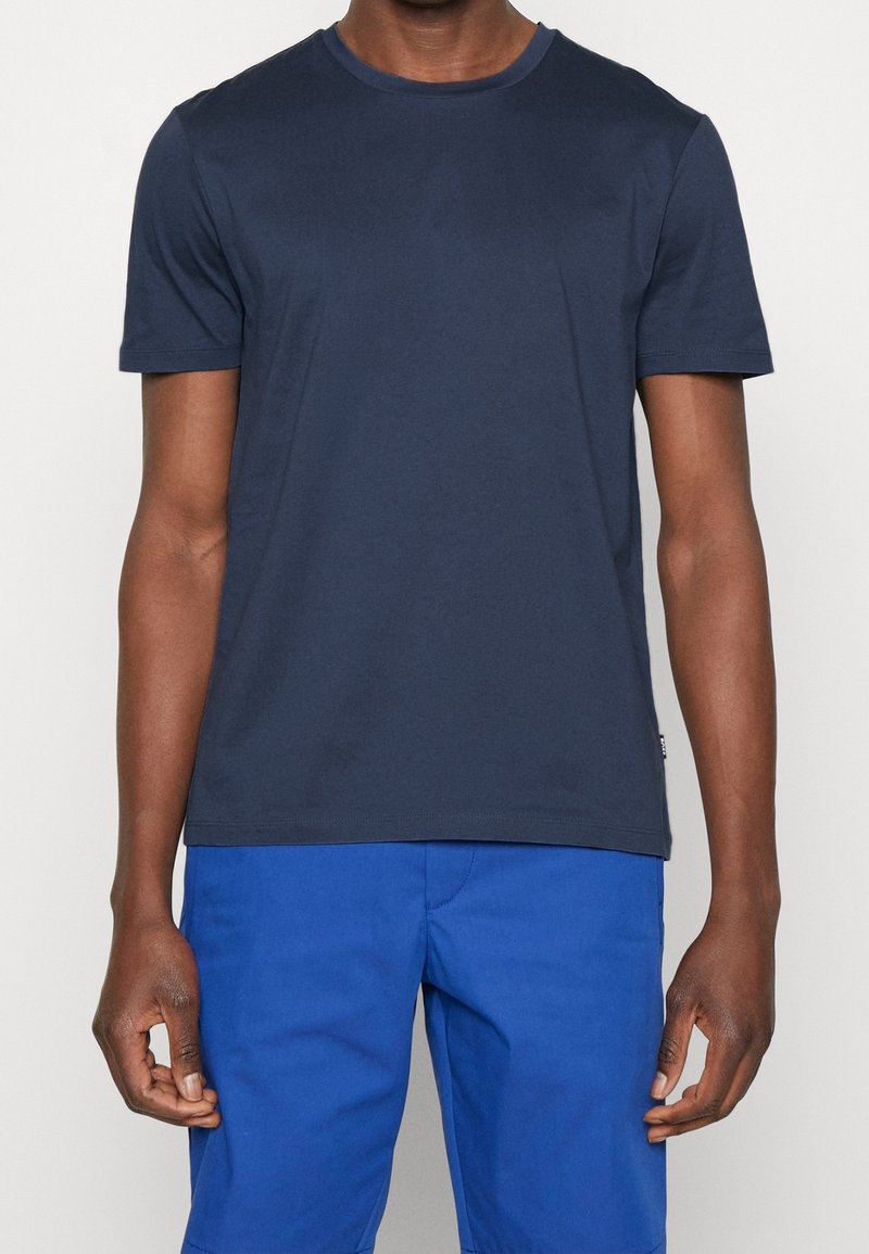 Homme portant un t-shirt à manches courtes bleu marine uni et un pantalon bleu vif, debout devant un fond clair uni.