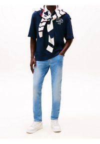 Polo de marina de manga corta con logo blanco, jeans de un azul claro y zapatillas blancas. Un suéter de punto a rayas está colocado sobre los hombros.