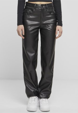 Henkilö pukeutuneena mustiin tekonahkahousuihin, joissa suorat lahkeet ja etutaskut, valkoiset tennarit jalassa, seisoo yksivärisen taustan edessä.