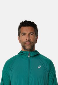 Veste légère à capuche couleur sarcelle avec fermeture éclair, présentant un petit logo sur la poitrine et un tissu texturé pour la respirabilité.