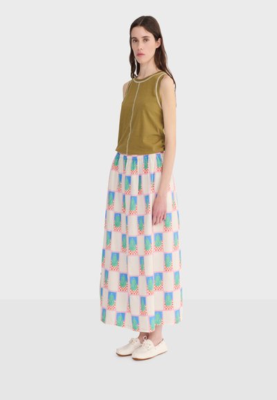 Žena v olivovo zelenej bezrukávovej blúzke a dlhej vzorovanej sukni s bielymi teniskami stojaca pred jednoduchým svetlým pozadím.