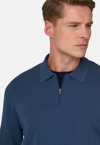 Marineblå polo-shirt med en tekstureret strik, med en lynlås i kraven og subtile syningsdetaljer, båret over et mørkt inderlag.