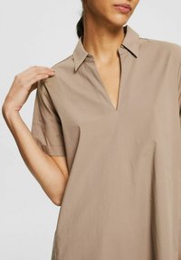 Beige kortärmad skjortklänning med v-ringning och krage. Tillverkad av ett mjukt tyg, med en avslappnad passform och subtil lyster.
