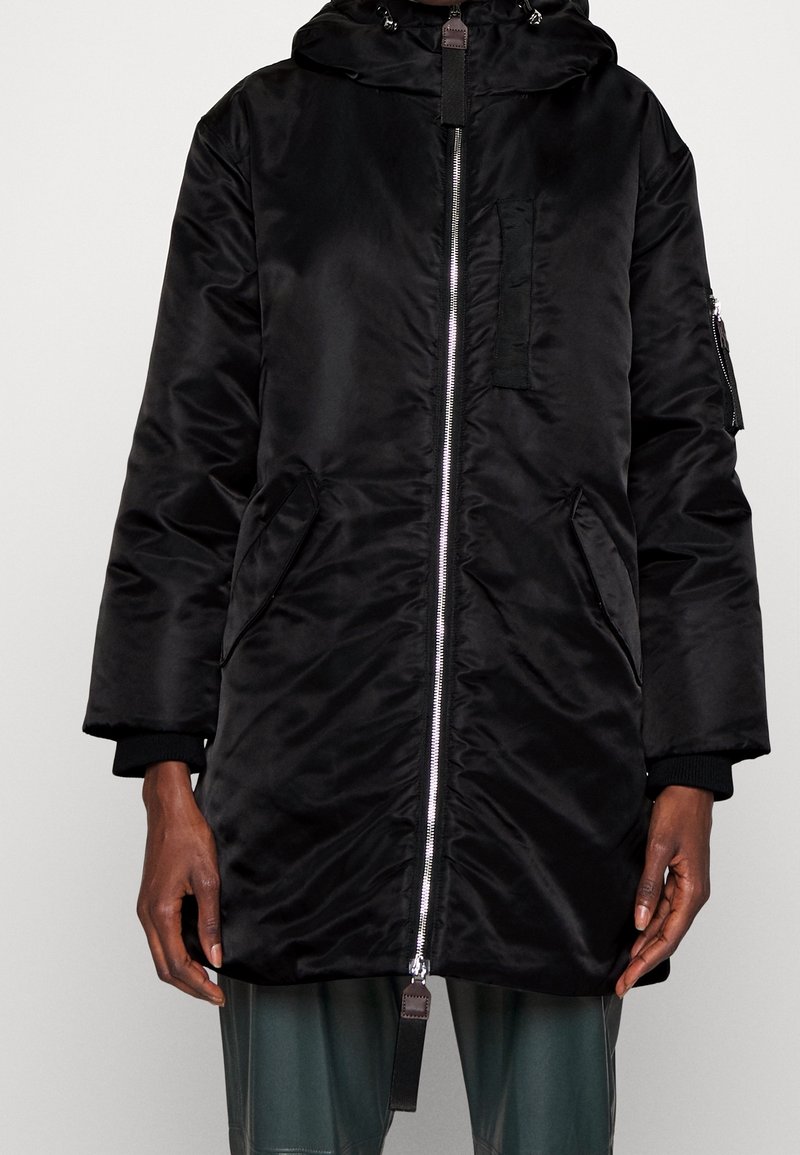 Giacca parka nera realizzata in un tessuto lucido e liscio. Dotata di cappuccio, cerniera frontale, tasche laterali e polsini a costine. Design minimalista.