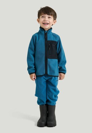 Ung dreng smiler og står iført en blå fleecejakke, matchende bukser og sorte gummistøvler på en ensfarvet baggrund.