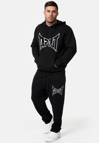 Conjunto de sudadera con capucha y pantalones de chándal en negro, hecho de una mezcla de algodón, con un prominente logo blanco de "TAPOUT". Cuerdas en la capucha, puños acanalados.
