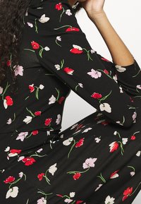 Kobieta w dopasowanej czarnej sukience z czerwonymi, białymi i różowymi kwiatowymi wzorami, siedząca z jedną ręką opartą na kolanie.