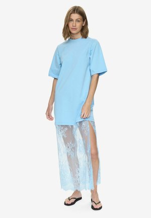 Kvinde iført en lys blå oversize t-shirtkjole med et gennemsigtigt blonder nederdelsoverlag og sorte klipklapper, stående mod en hvid baggrund.