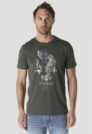 Hombre con camiseta verde oscuro con un gráfico abstracto de calavera y vaqueros azul claro, de pie frente a un fondo gris liso.