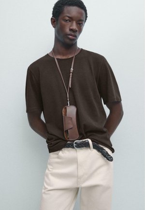 Junger Mann, der ein dunkelbraunes T-Shirt, beige Hose mit einem geflochtenen schwarzen Gürtel und eine braune Ledertasche trägt, die an einem Riemen um seinen Hals hängt.