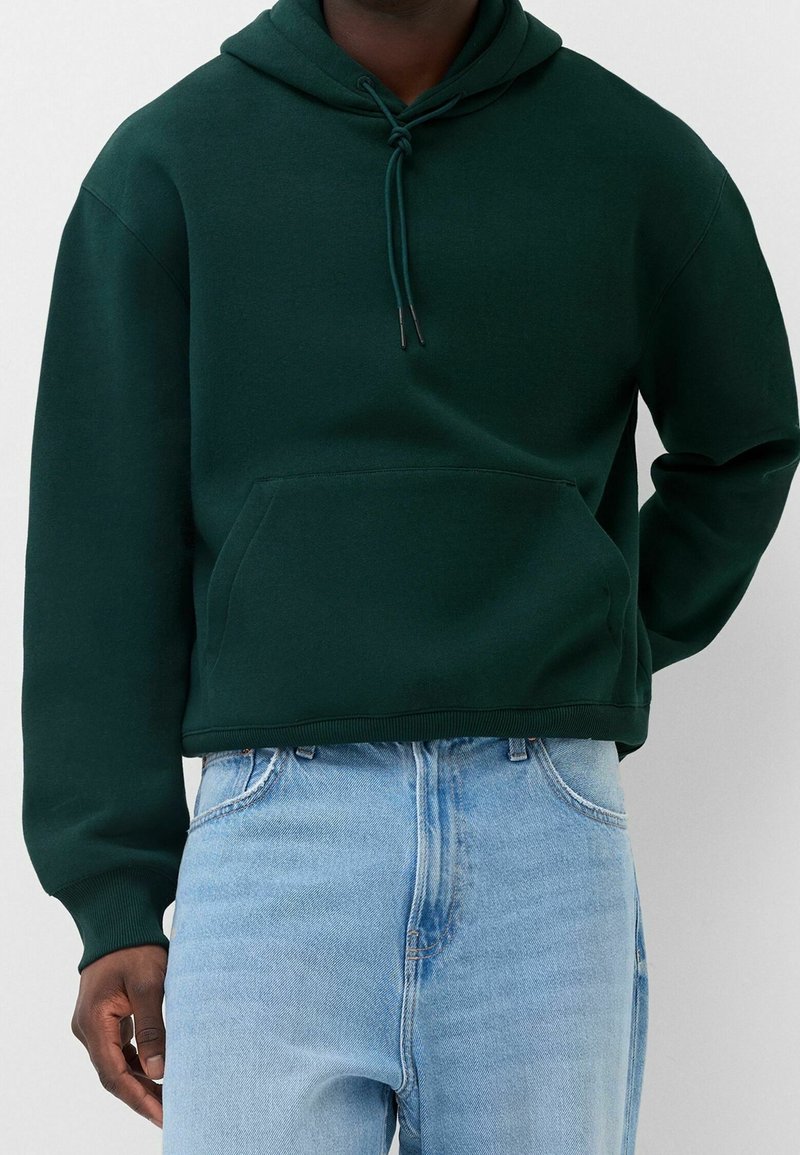 Personne portant un sweat à capuche vert foncé avec une poche devant et un jean taille haute bleu clair sur un fond uni.