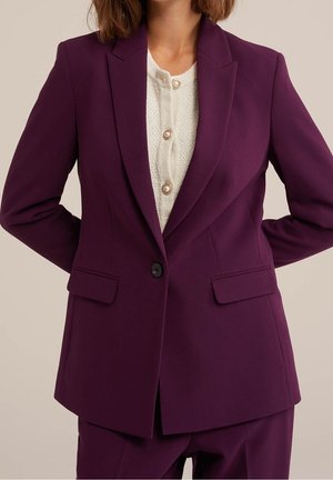 Femme portant un blazer violet foncé sur mesure par-dessus un chemisier blanc texturé avec des boutons en perle, debout, les mains dans le dos.