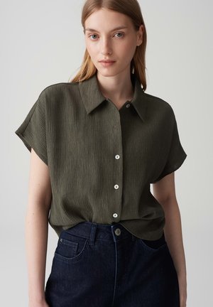 Olive green, short-sleeve blouse with a textured fabric, a collared neckline, and white buttons, paired with dark denim jeans.