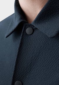 Abrigo texturizado en azul marino con cuello y botones de presión negros. La tela presenta un patrón sutil y un acabado suave.