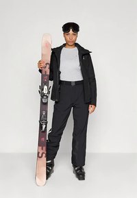 Sprzęt narciarski: czarna kurtka, szara koszula, czarne spodnie, buty narciarskie. Trzyma różowe i brązowe narty z wiązaniami, na głowie ciemne gogle narciarskie.