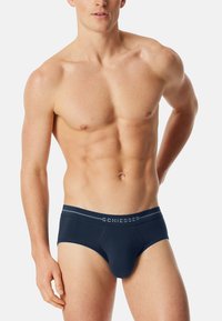 Kaal bovenlijf mannelijke model in marineblauwe Schiesser onderbroek, staand met één arm achter zijn rug tegen een plain witte achtergrond.