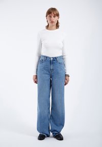 High-waisted blue denim jeans with wide legs, featuring a button fly, five pockets, and light fading, paired with a white long-sleeve shirt.