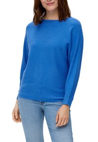 Blå ribbad tröja med vid halsringning och långa ärmar, med ett strukturerat mönster och en figurnära nederkant. Kombinerad med ljusa jeans.