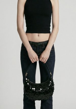 Personne portant un haut noir sans manches et un jean foncé tient un sac à main noir pailleté par les poignées sur un fond uni.