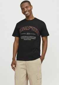 Zwarte katoenen T-shirt met witte en oranje bedrukte tekst, korte mouwen en een ronde hals. G gedragen met beige cargobroek.