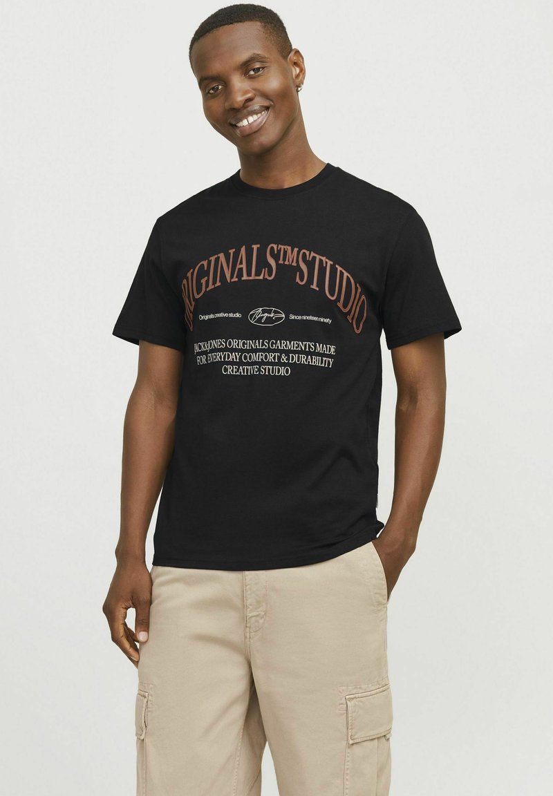 Zwarte katoenen T-shirt met witte en oranje bedrukte tekst, korte mouwen en een ronde hals. G gedragen met beige cargobroek.