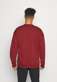 Uomo con capelli ricci corti che indossa una felpa rossa semplice e pantaloni neri, in piedi di fronte a un muro bianco.