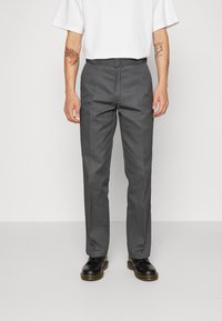 Grijze broek van geweven stof, met een rechte snit, frontzakken en contrasterende zijpiping. Draagt met zwarte schoenen.