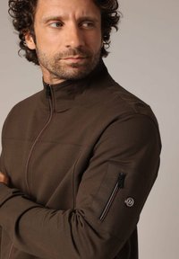 Veste zippée marron avec un col haut, présentant des coutures d'accent rouges et une poche zippée sur le bras gauche. Tissu doux et texturé.