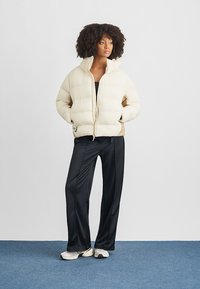 Ženska stoji z rokami v žepih, oblečena v kremno puhovko, črne široke hlače in bele superge na modrem preproganem tleh.