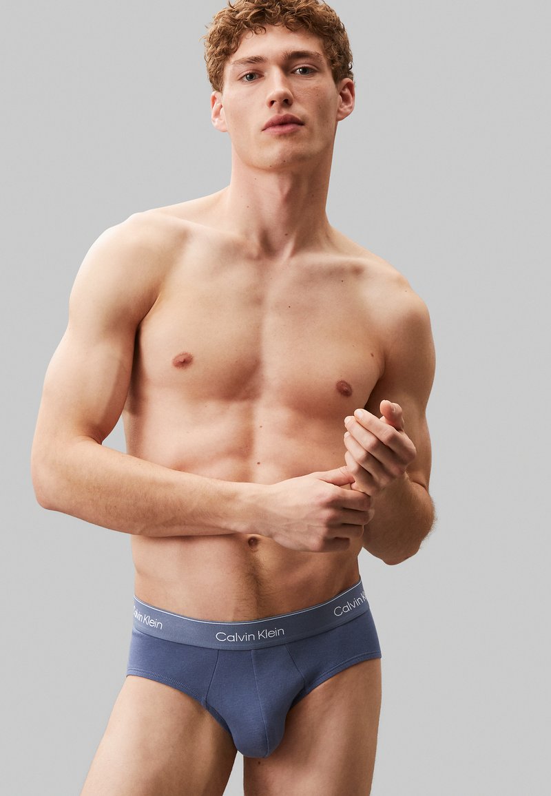 Slip in cotone blu con una morbida fascia in vita che presenta il marchio "Calvin Klein". La vestibilità è aderente, evidenziando la fisicità del modello.