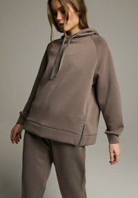 Brązowa bluza z kapturem o luźnym kroju, z ściśniętymi mankietami, kieszenią kangurką i bocznymi detalami. Miękka faktura.