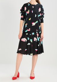 Zwarte sweatshirt met kleurrijke, abstracte vormen, gecombineerd met een geplooide zwarte rok met soortgelijke patronen. Model draagt rode hoge hakken.